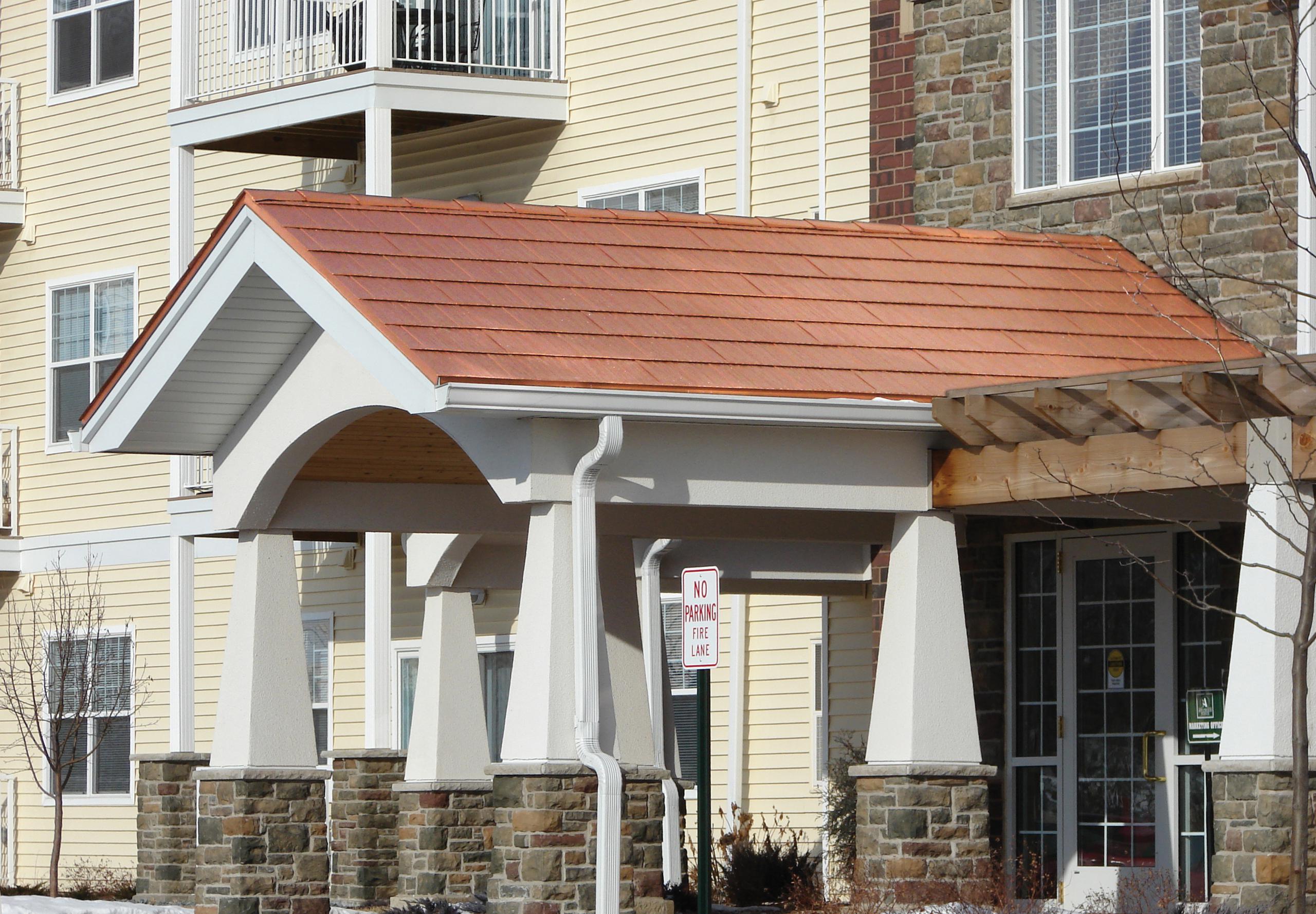The covering of the entrance to an apartment complex showcases the Arrowline Shake Copper Roofing to bring out the rich colors of the stone and brick installed on the building.